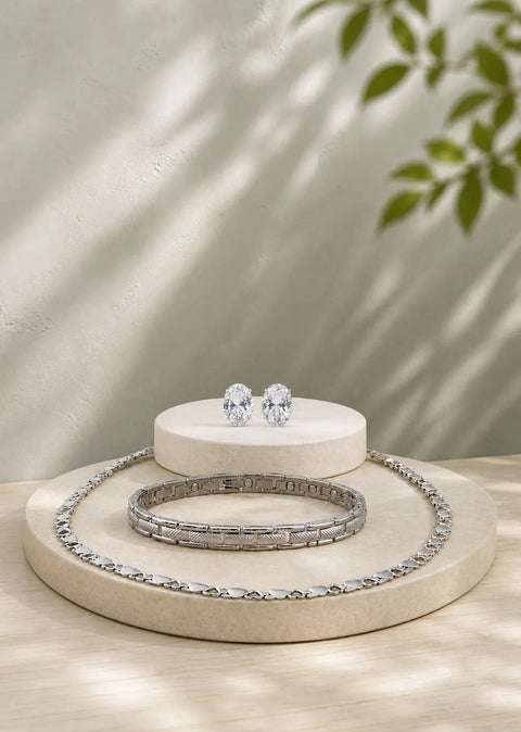 A silver-toned jewelry set displayed on round beige pedestals, including a tennis-style necklace, a bangle bracelet, and a pair of stud earrings with clear gemstones, set against a softly lit wall with leaf shadows.