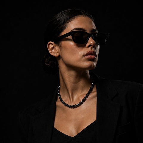 A woman in a black blazer and black top wearing black cat-eye sunglasses and a black beaded necklace, photographed against a solid black background.