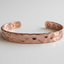 A hammered copper cuff bracelet with a hammered texture and circular cutouts on the ends, displayed on a light gray background.