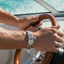 A close-up lifestyle shot of a man's hands steering a boat, with one hand wearing a silver-toned leather bracelet featuring gold-toned circular accents.